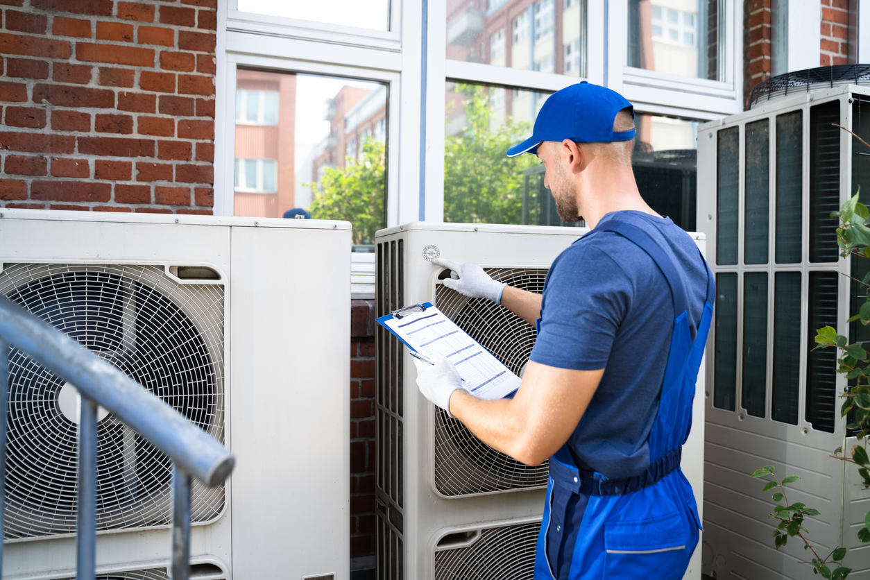 Schedule Servicing: A technician in blue overalls inspects outdoor air conditioning units and takes notes on a clipboard.